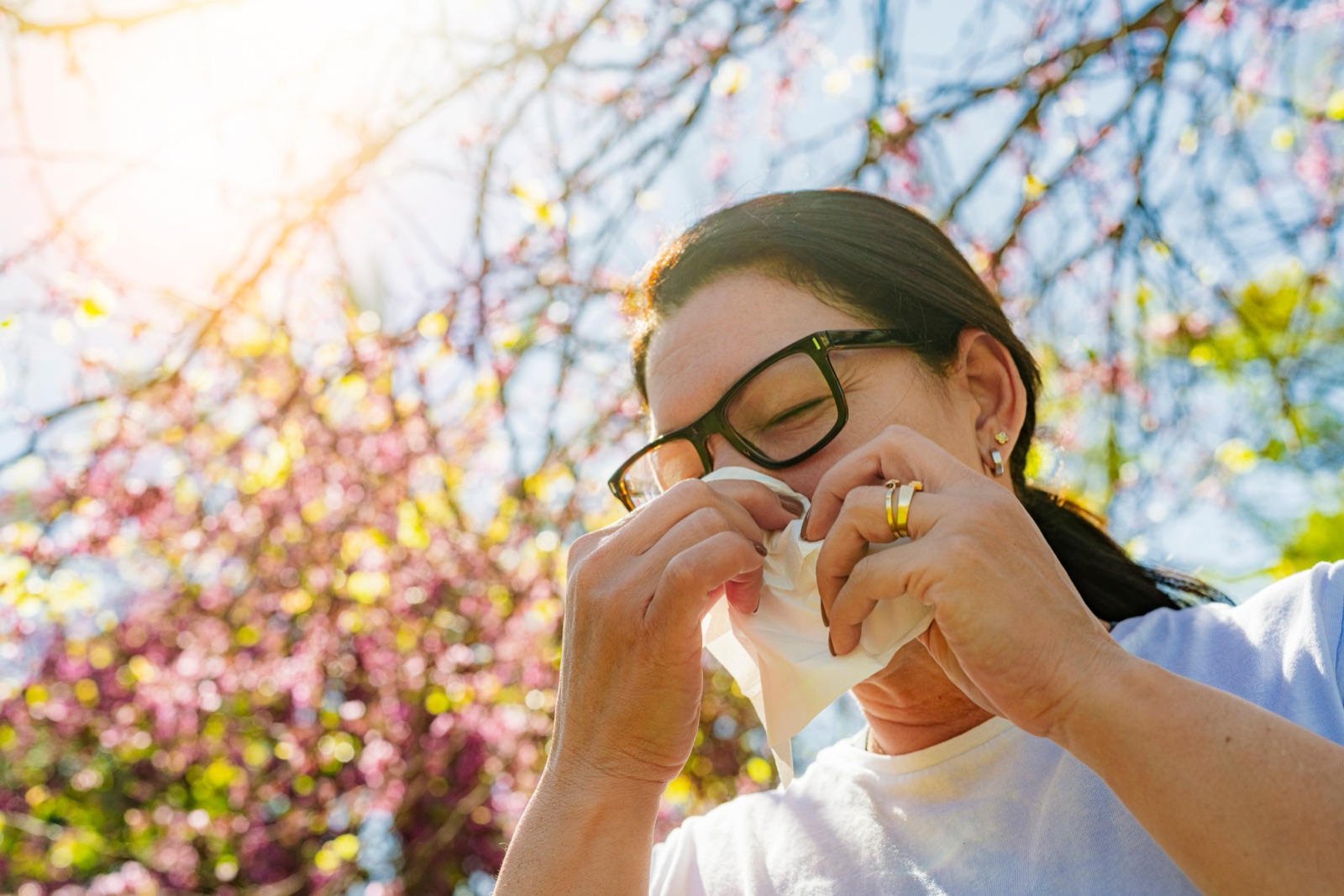 Woman in glasses holding a tissue to her nose beneath a tree in spring blossom