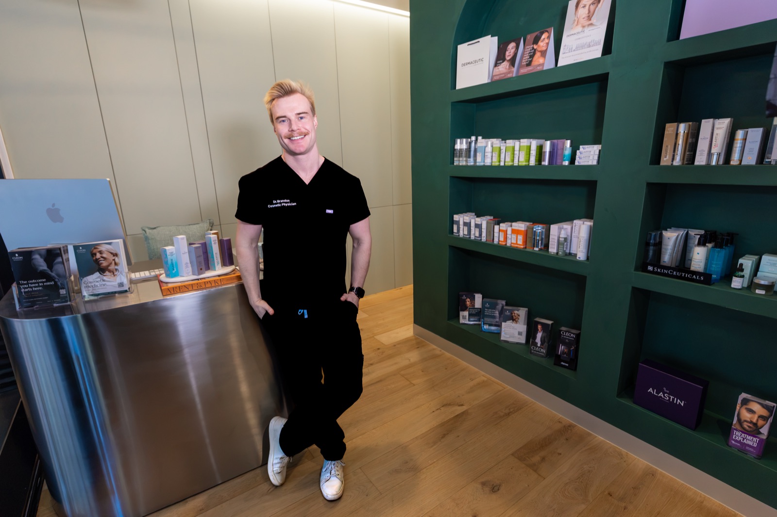 Dr Brandon smiling beside the front desk and the green product shelves in the Cleon clinic in Potts Point, Sydney