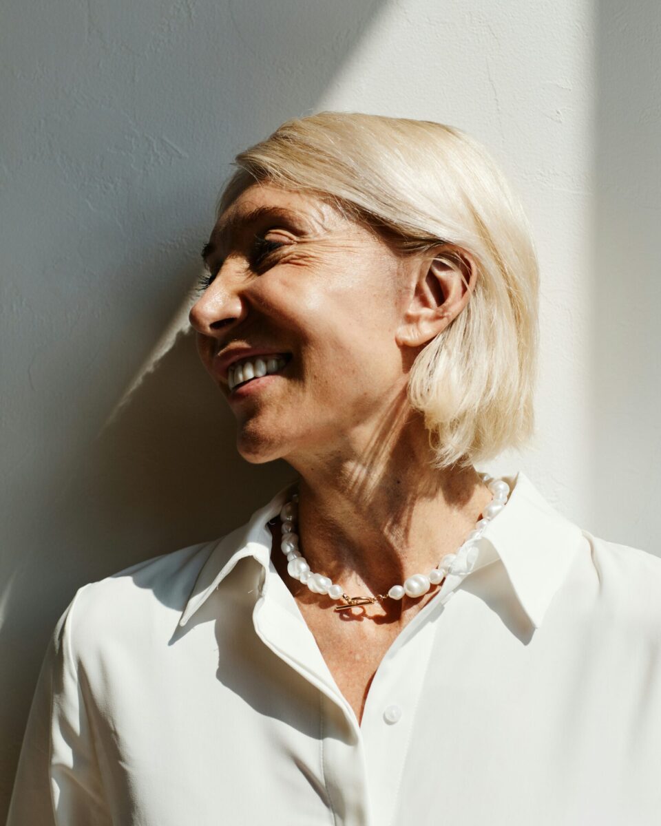 Smiling woman with blonde hair in a white shirt and pearl necklace against a Cleon Clinic branded backdrop