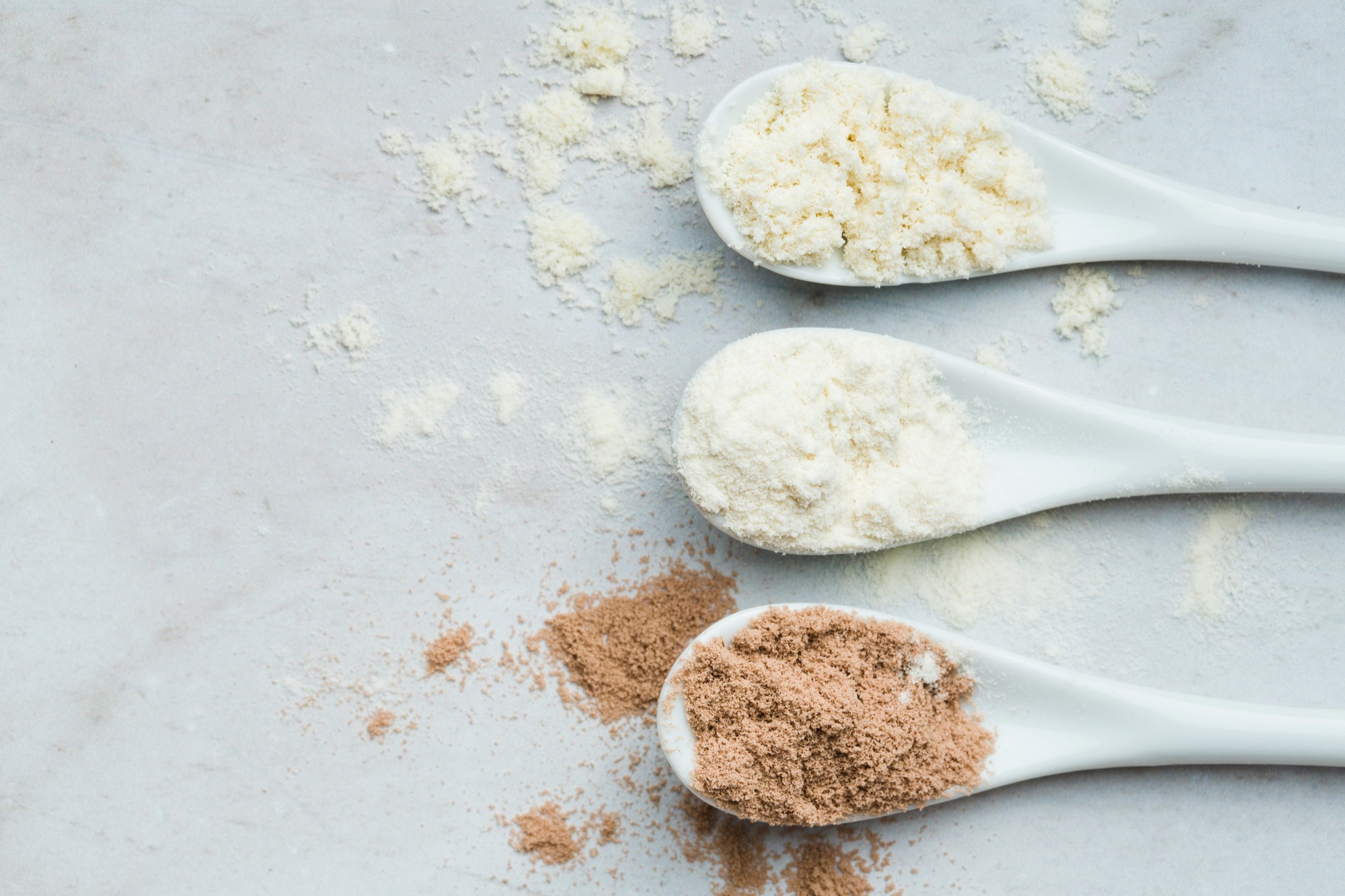 Three white spoons of collagen supplement powder, two cream and one cocoa, on a pale marble surface